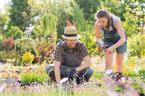 A garden scene showing two women working together in a landscaped outdoor space. The woman in a straw hat and dark apron is kneeling on a lush green lawn, carefully examining or tending to flowering plants with small purple and pink blossoms. The other woman, with long dark hair and denim overalls, is standing nearby, leaning forward to assist or observe, holding garden shears. The garden features a variety of colorful flower beds, bordered by soil and low hedges, with a backdrop of tall, leafy trees and shrubs. The scene is well-lit with natural daylight, indicating a bright and pleasant day in a suburban garden in Parsons Green, London. The environment includes neatly maintained grass, flower beds with a diverse mix of plants, and a clean, well-organized paved area, reflecting professional gardening and lawn care services by Lawn Mowing Parsons Green.